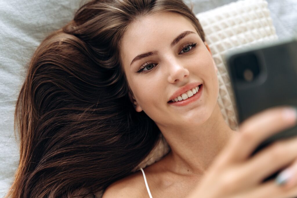 Delighted woman using smartphone in bed. Young smiling woman laying on the bed at home