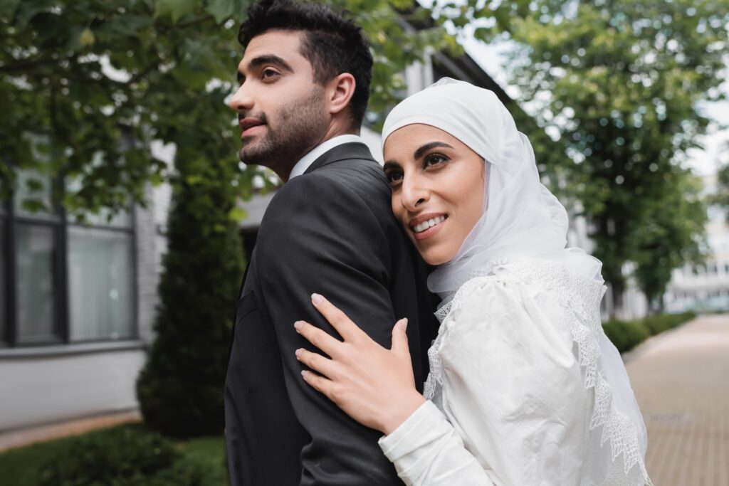 pleased muslim bride in hijab and wedding dress hugging groom